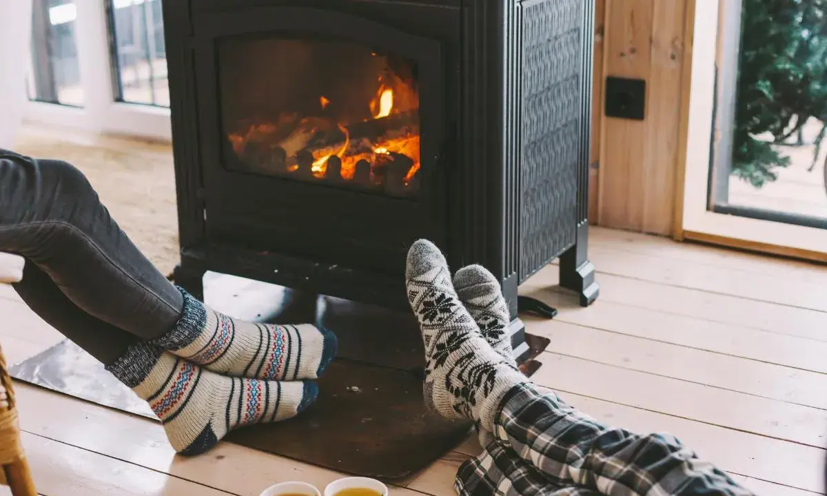 Pies con calcetines calentándose junto a una estufa de leña, disfrutando de la calefacción sin electricidad y una taza de té.