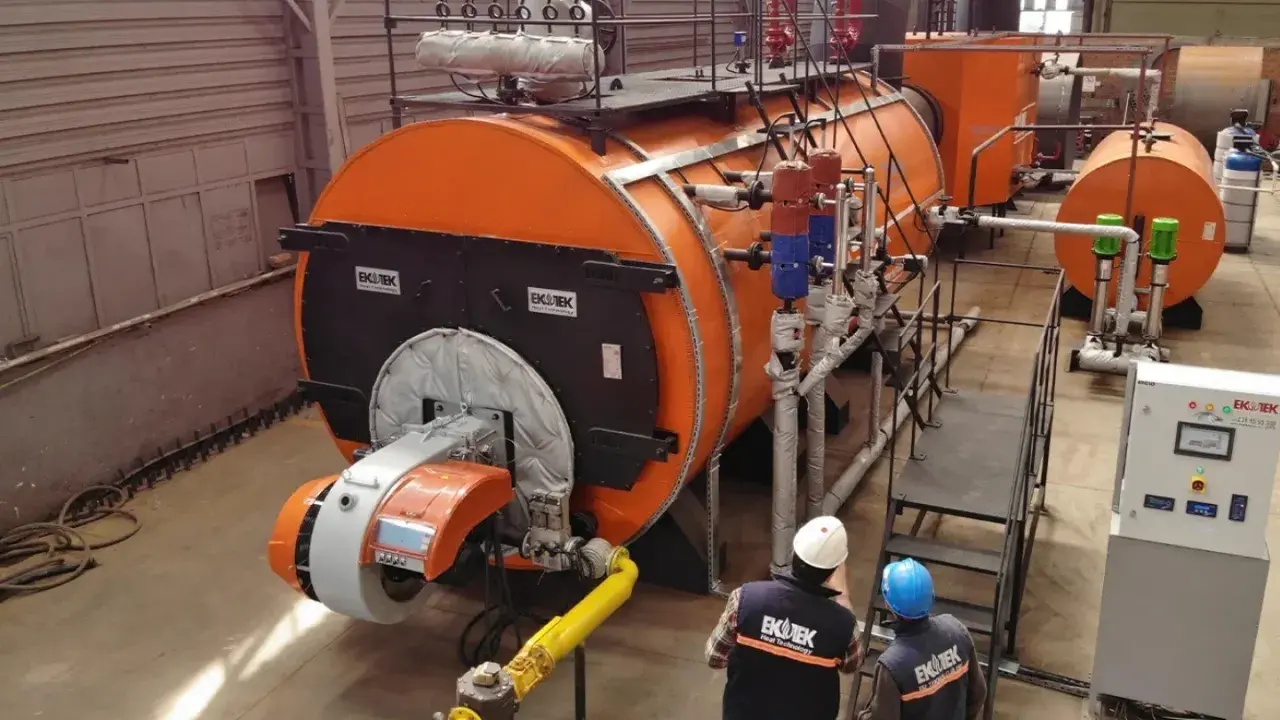 Dos técnicos observan varios tipos de calderas industriales naranjas y grises en una fábrica.