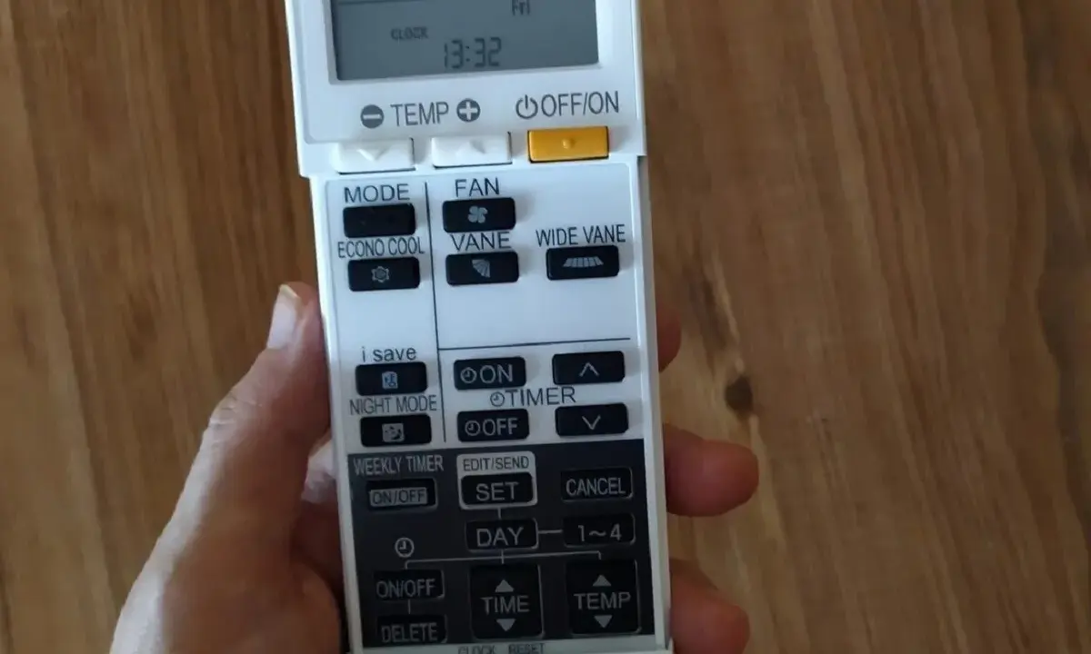 Mano sujetando un control remoto de aire acondicionado. Se ven botones como "MODE", "FAN", "ECONO COOL", "VANE", "WIDE VANE", "NIGHT MODE", "TIMER", "WEEKLY TIMER", "SET", "DAY", "TIME", "TEMP" y "DELETE".