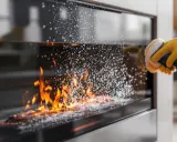 Mano con guante amarillo limpiando el cristal de la chimenea con un cepillo eléctrico. Las llamas arden detrás del cristal cubierto de burbujas.