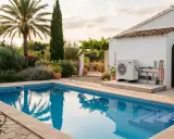 Piscina de baldosas azules con escalones, rodeada de un jardín exuberante y una casa blanca. El sol del atardecer ilumina el **suelo radiante** de la zona de la **piscina**.