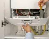 Mano ajustando una caldera de gas moderna, no una de las calderas de gas antiguas. Se ven tuberías de cobre y componentes internos.