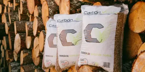 Bolsas de pellets de madera "Carbón Verde" apiladas contra troncos. Si tu estufa de pellets muestra un error 3, revisa la carga de combustible.