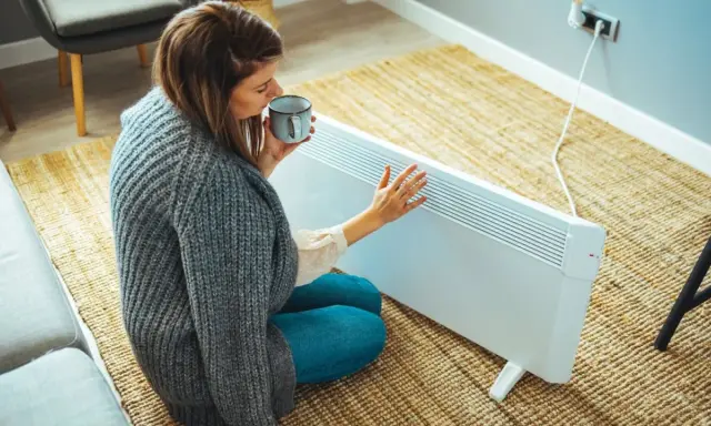 Mujer con taza de té cerca de un calefactor eléctrico, una solución para la alternativa al gas en un piso.