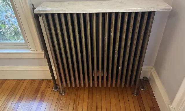 Radiador de latón con tapa de mármol, un ejemplo de calefacción antigua, junto a una ventana.
