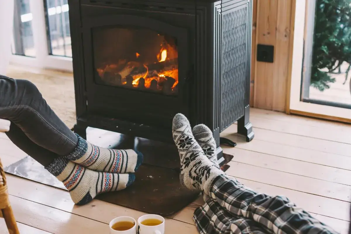 Pies con calcetines calentándose junto a una estufa de leña, disfrutando de la calefacción sin electricidad y una taza de té.