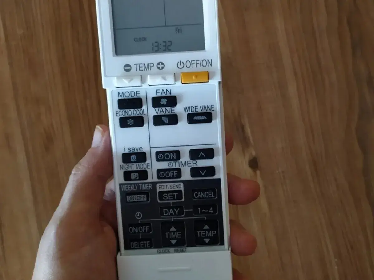 Mano sujetando un control remoto de aire acondicionado. Se ven botones como "MODE", "FAN", "ECONO COOL", "VANE", "WIDE VANE", "NIGHT MODE", "TIMER", "WEEKLY TIMER", "SET", "DAY", "TIME", "TEMP" y "DELETE".