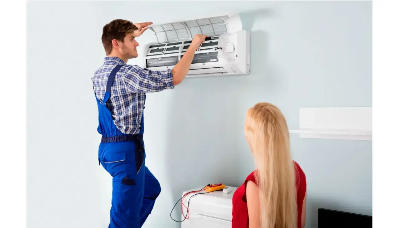 T&eacute;cnico realiza mantenimiento preventivo de aire acondicionado mientras una mujer observa.